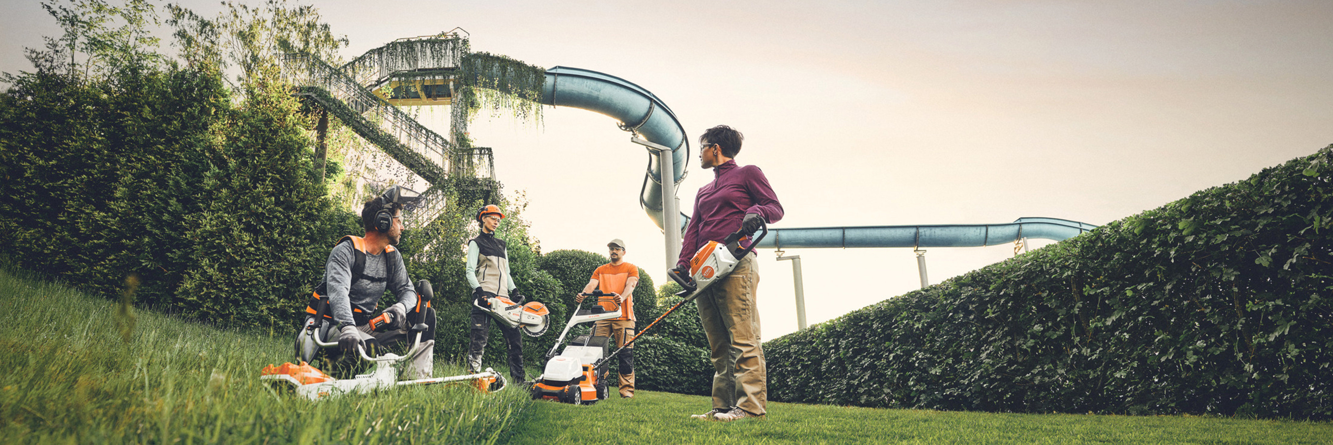 In einer Grünanlage mit einer großen Hecke und Wasserrutschen im Hintergrund arbeiten mehrere Personen mit STIHL Gartengeräten. Sie sind mit Freischneidern, Rasenmähern und Schutzausrüstung ausgestattet.