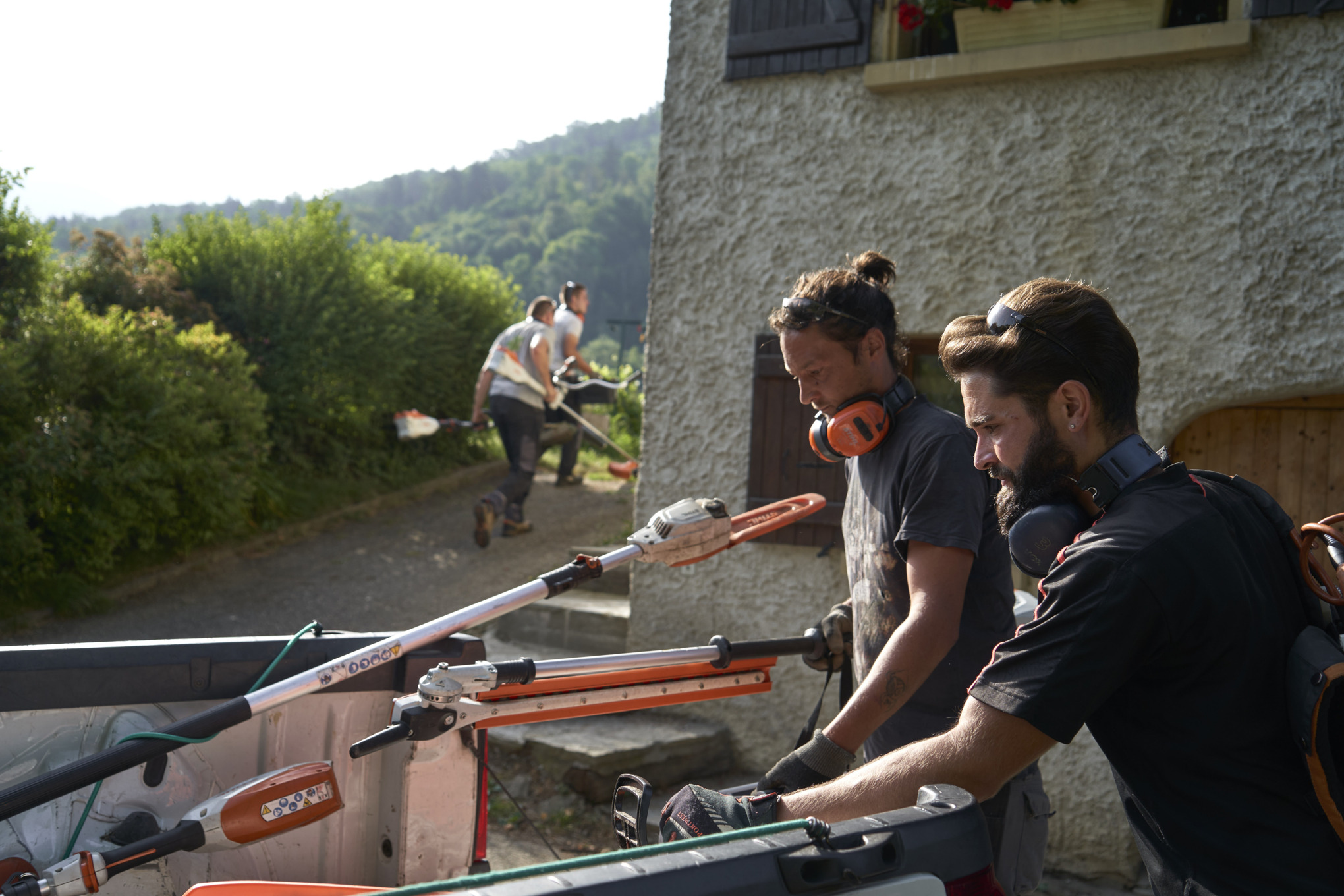 Zwei Landschaftsgärtner entladen STIHL Akku-Geräte aus einem Auto, im Hintergrund sind weitere Arbeiter