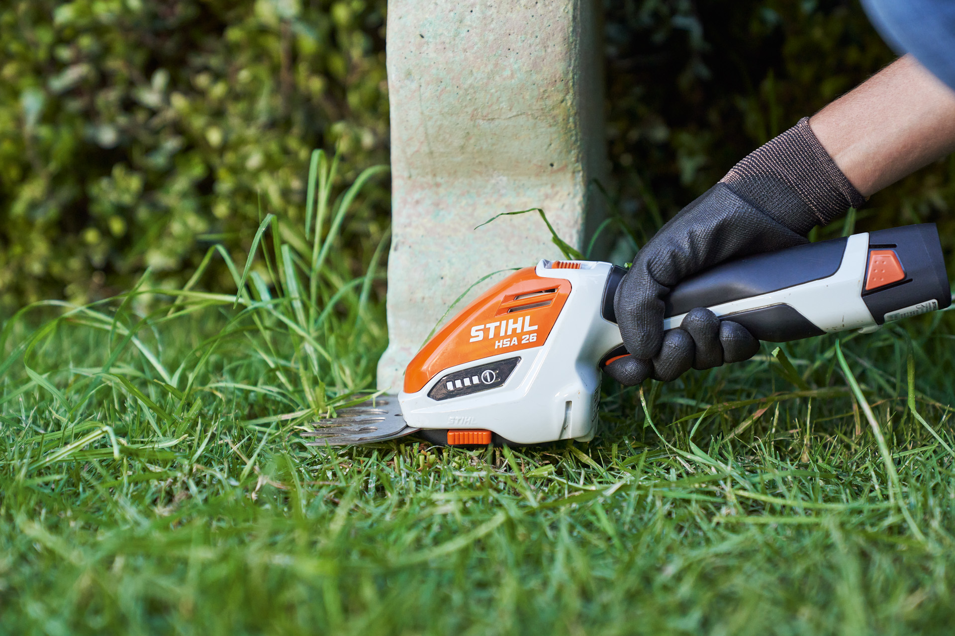 Nahaufnahme: Mit Schutzhandschuh versehene Hand trimmt mit STIHL Akku-Strauchschere HSA 26 Rasenkante um Bank herum