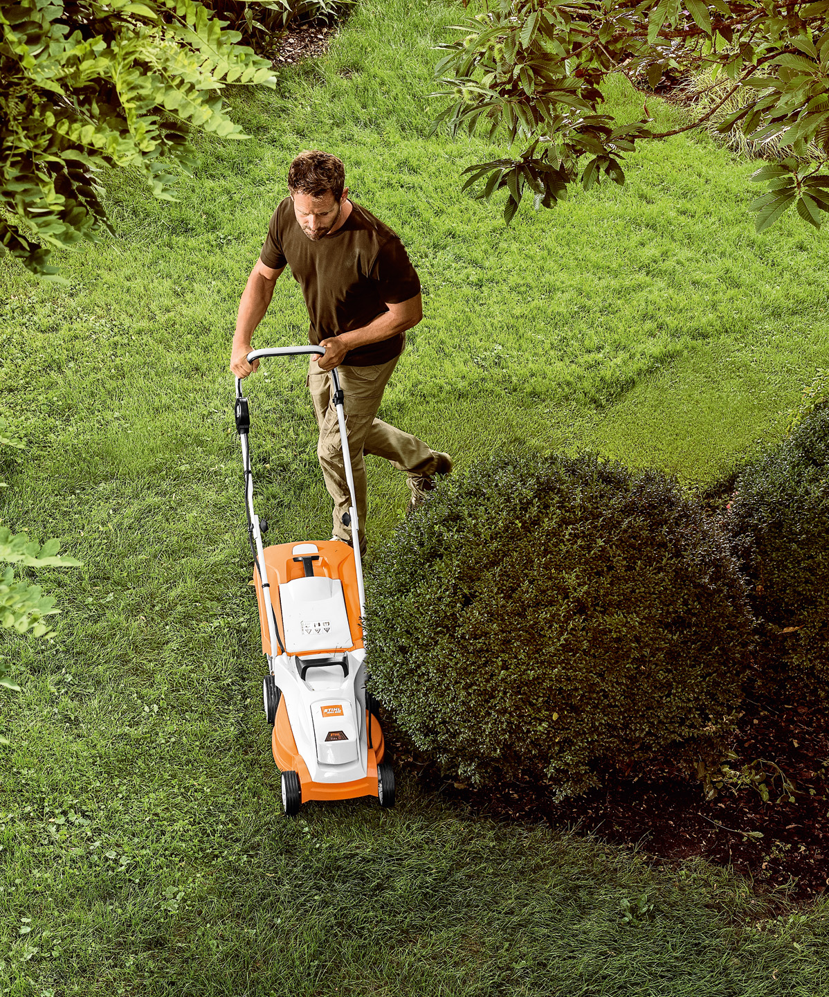 Blick von oben: Mann mäht eine Rasenfläche in einem kleinen Garten mit STIHL Akku-Rasenmäher RMA 235 rund um kleinen Baum herum.