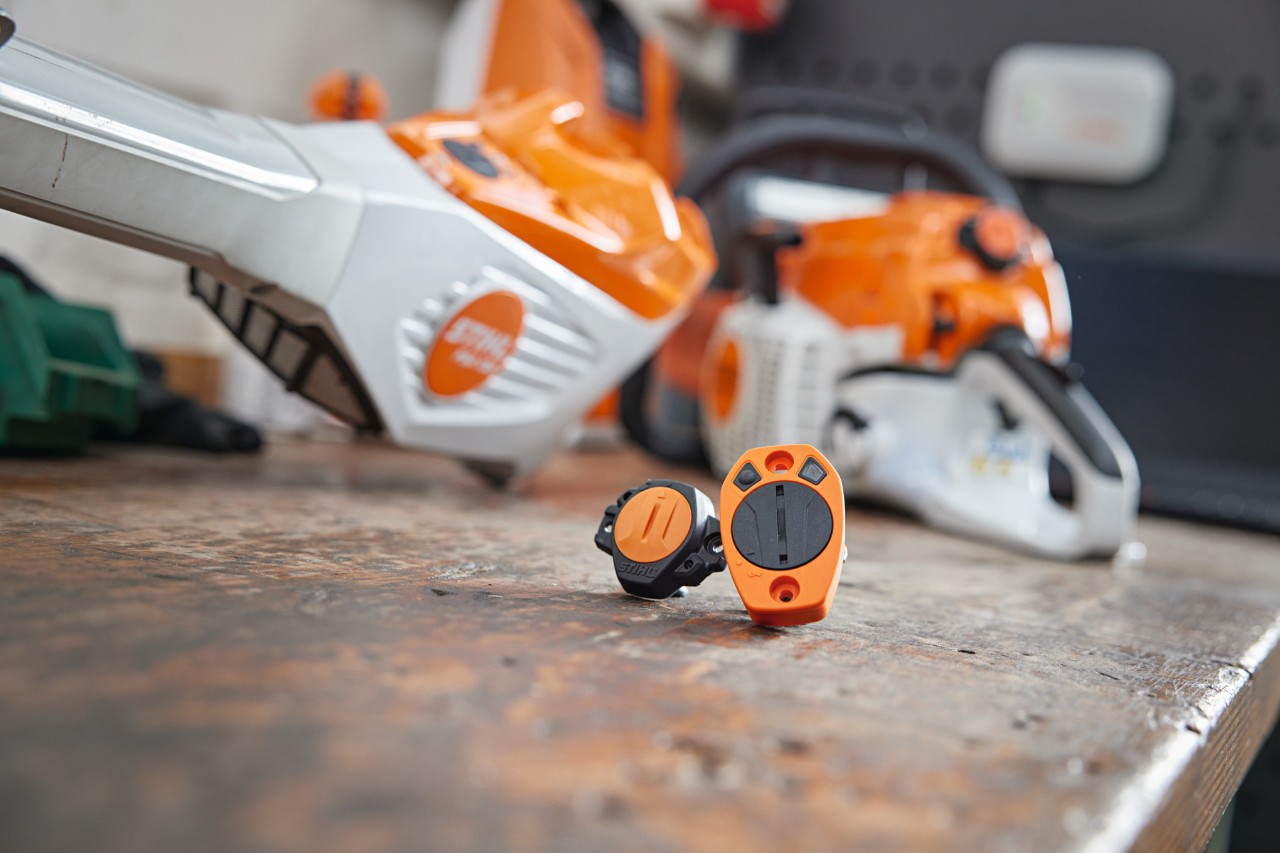 Ein STIHL Smart Connector und ein STIHL Smart Connector 2 A liegen auf einer Werkbank. Im Hintergrund sind in Unschärfe verschiedene STIHL Geräte zu sehen.