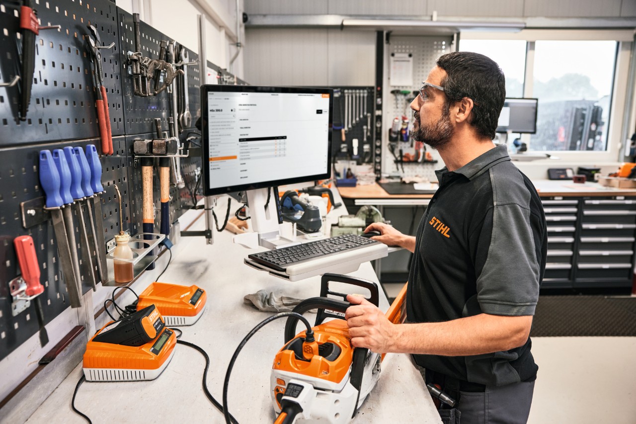 In einer Werkstatt steht im Vordergrund ein Mann, der einen PC bedient. Auf dem Tisch vor sich liegt eine Motorsäge