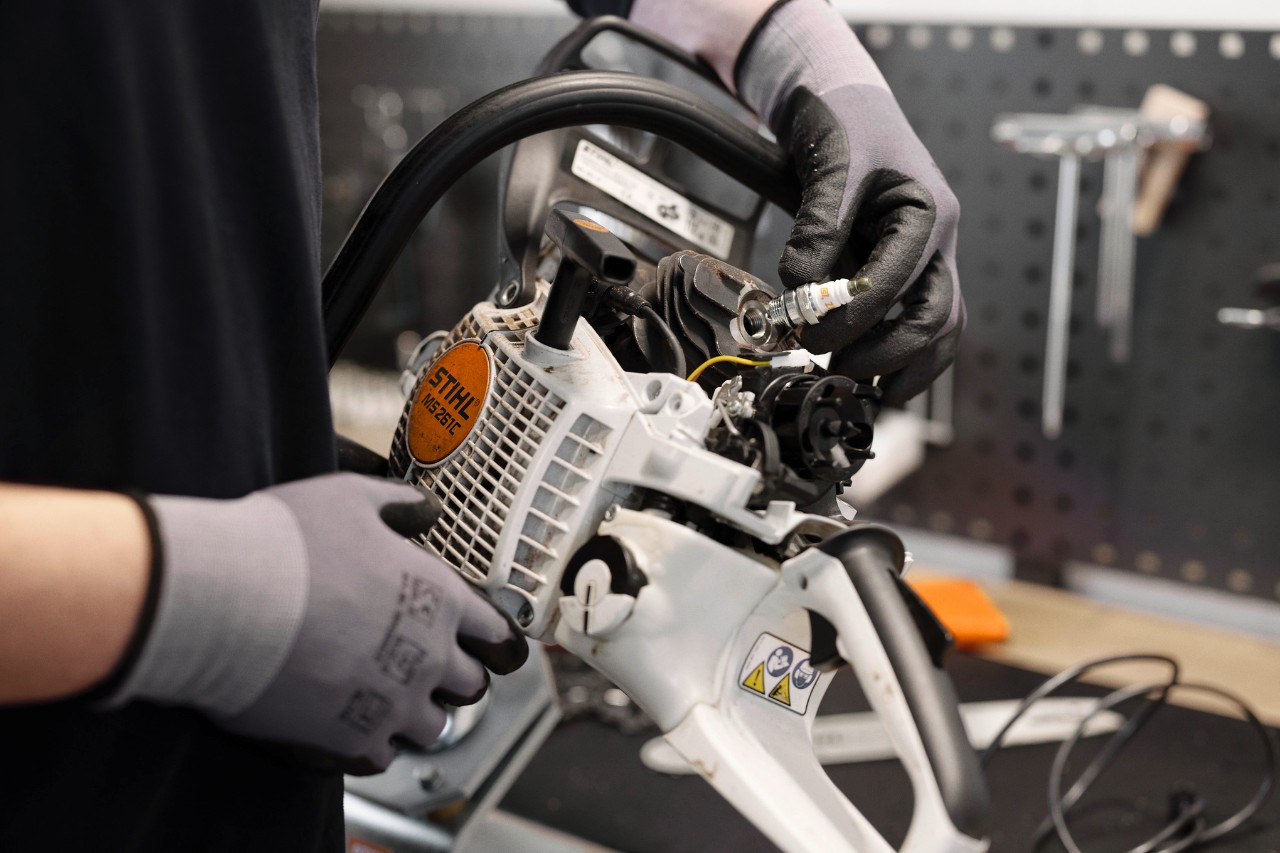 In einer Werkstatt steht im Vordergrund eine Person mit Handschuhen, die den Motor einer Motorsäge in den Händen hält und eine Zündkerze wechselt.