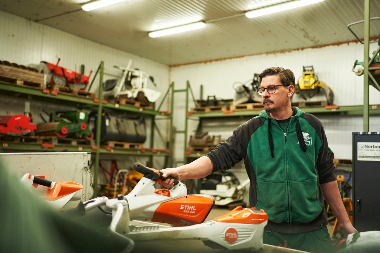 Ein Mann in einer Werkstatt steht hinter einer Arbeitsfläche mit STIHL Akku-Geräten, er greift nach dem Akku-Laubbläser STIHL BGA 200