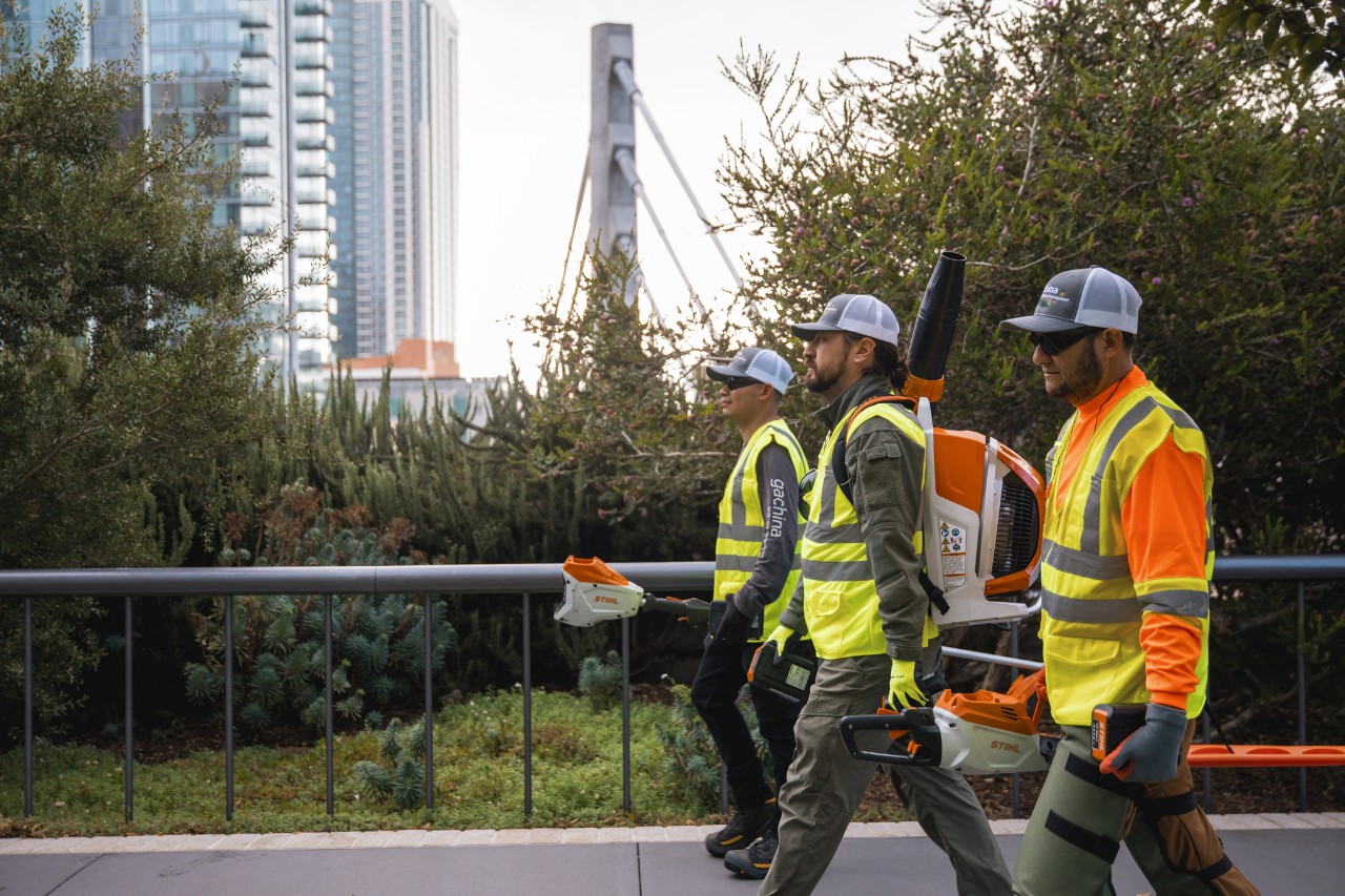 Drei LaGa-Arbeiter laufen mit Arbeitskleidung und STIHL Geräten durch eine urbane City.