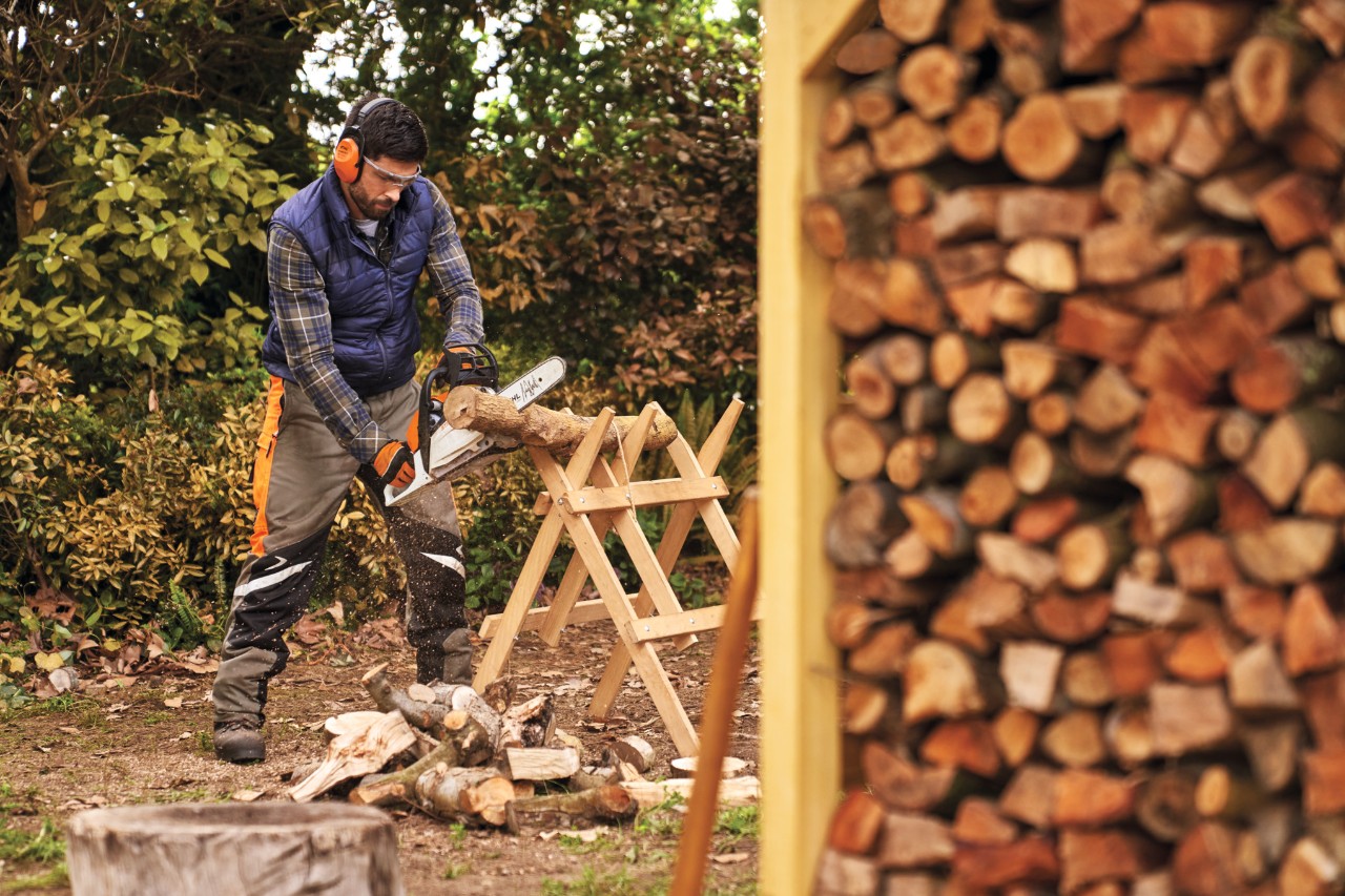 Mann in Schutzausrüstung sägt Holzscheite für den Brennholzunterstand zurecht