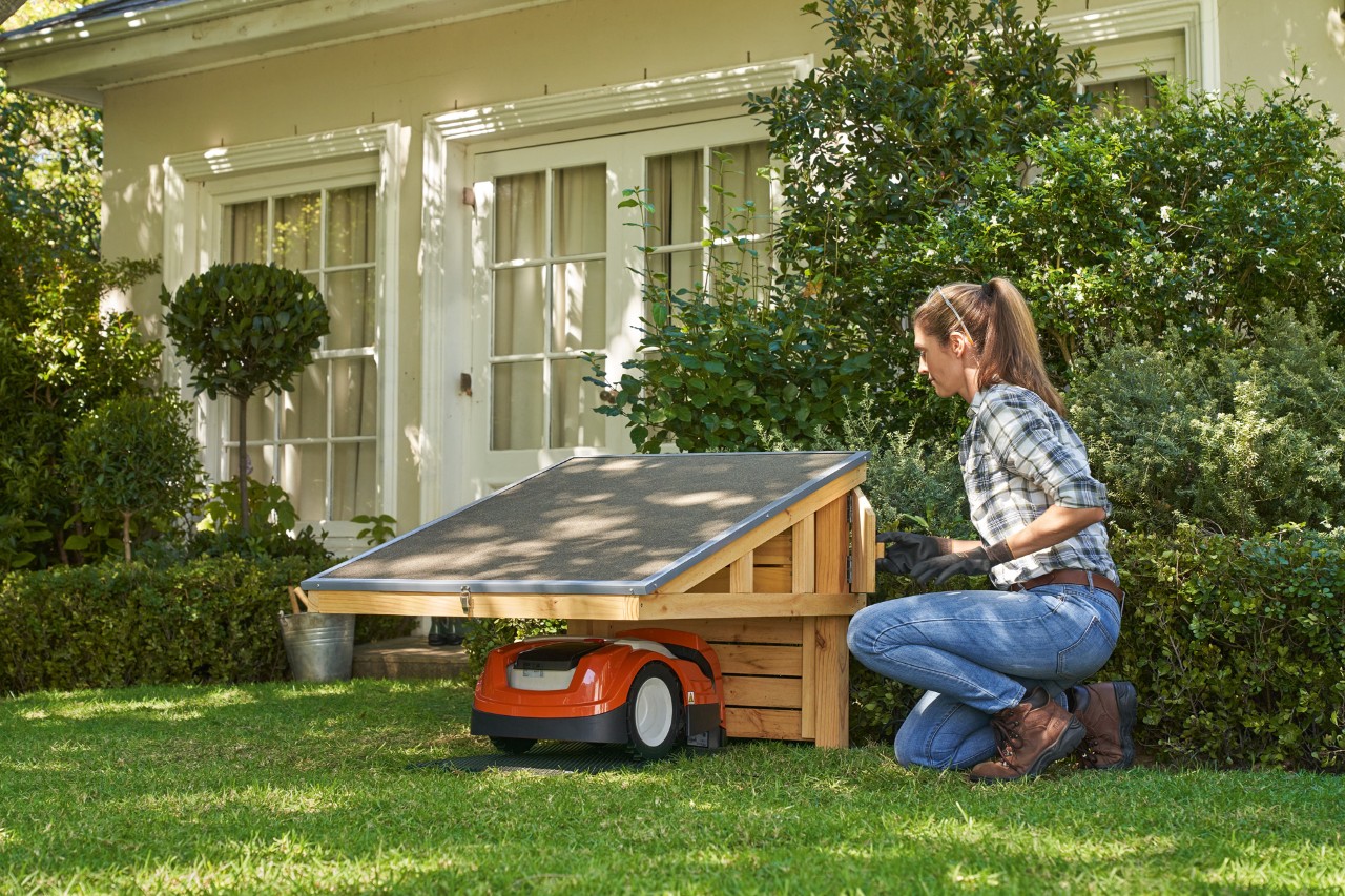 Frau mit Schutzbrille und Arbeitshandschuhen kniet auf Rasen neben  STIHL iMOW®-Garage mit geparktem STIHL iMOW® Mähroboter, öffnet Ablagefach    