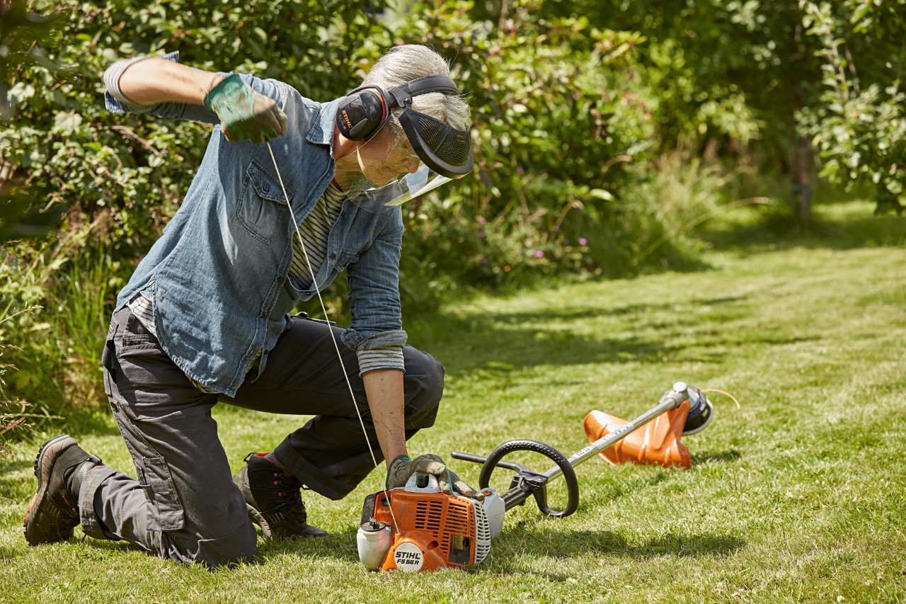 Frau mit Gehör- und Gesichtsschutz ist dabei, auf einem grünen Grundstück eine STIHL Benzin-Motorsense FS 56 R zu starten