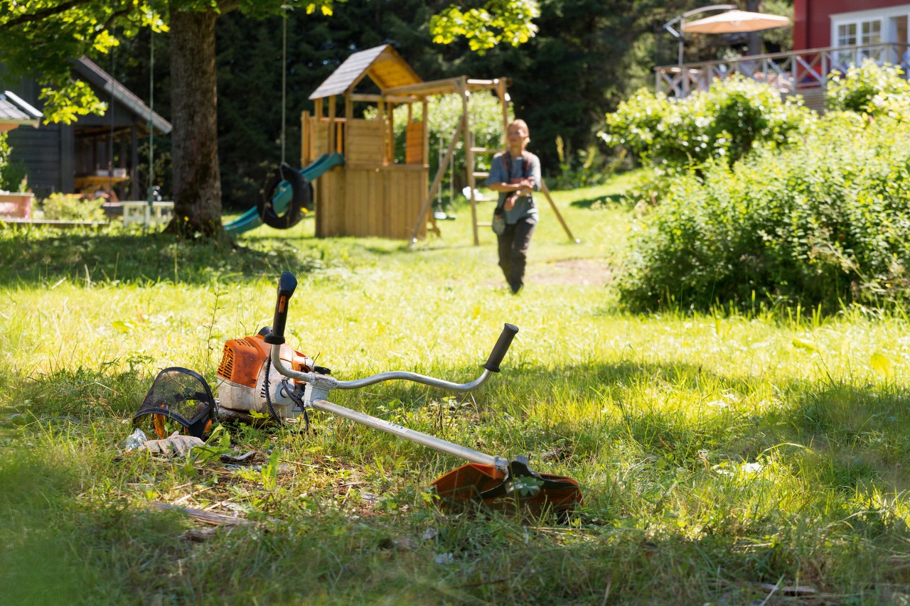Motorsense STIHL FS 240 liegt im Gras, im Hintergrund bereitet ein Mann sich auf die Wartung vor