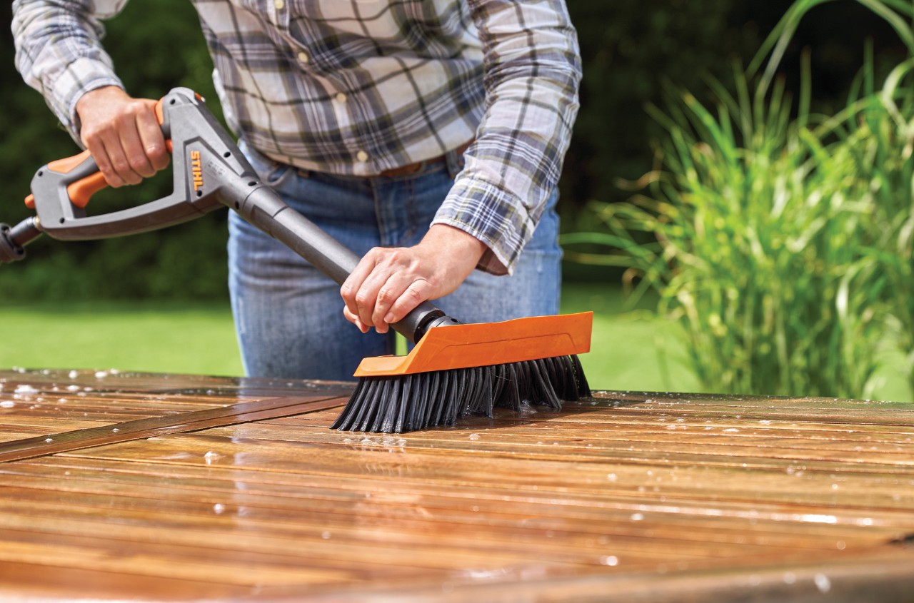 Frau reinigt Gartentisch aus Holz mit einem STIHL Hochdruckreiniger RE 130 plus mit passender Waschbürste