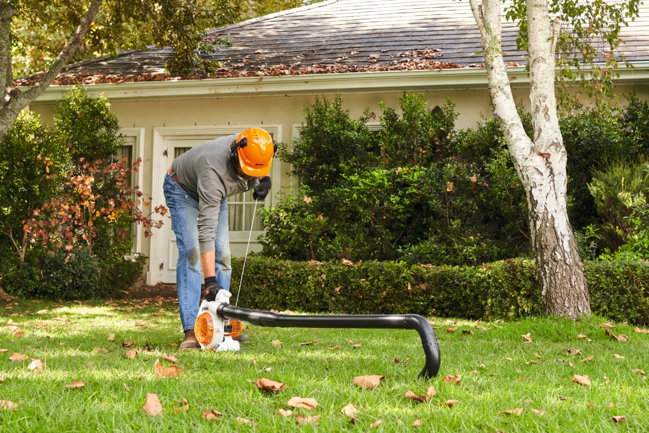 Ein Mann mit Helm und Schutzhandschuhen auf einem Gartengrundstück beugt sich über ein Blasgerät, um es anzuschalten
