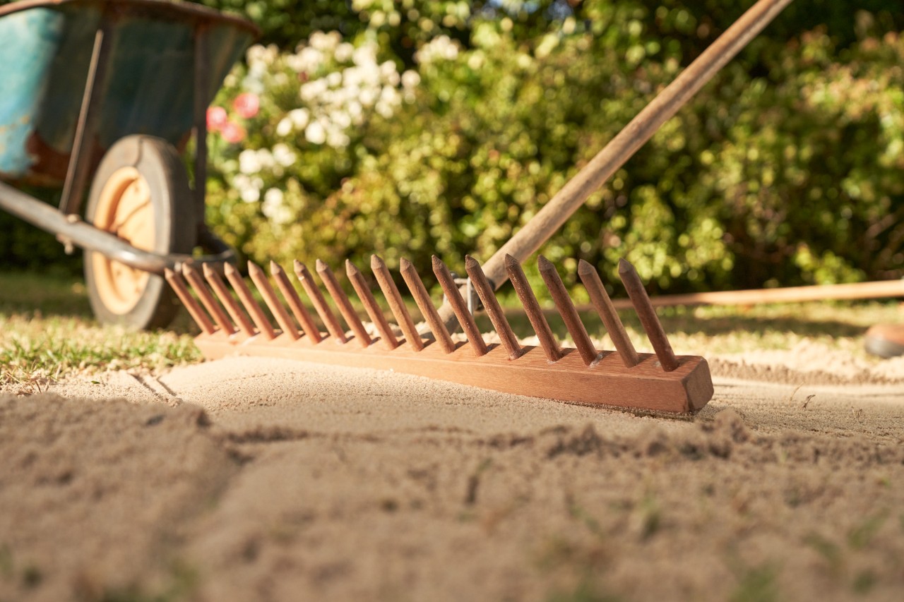 Mit der Rückseite einer Harke wird Sand auf einer ebenen Fläche geglättet, im Hintergrund Schubkarre und Hecke