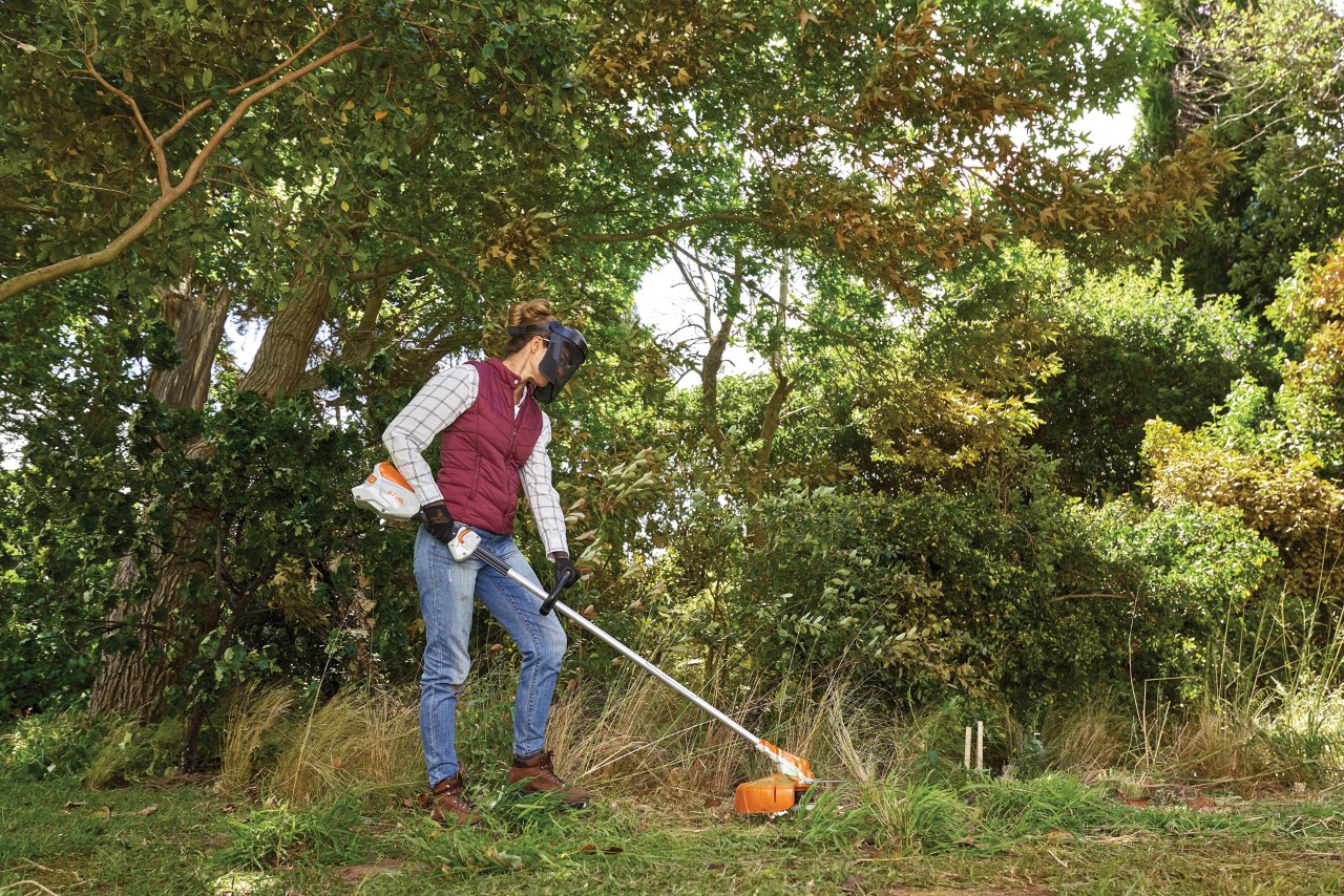 Eine Frau mit Gesichtsschutz mäht mit einem STIHL Freischneider den Rasen im verwilderten Garten