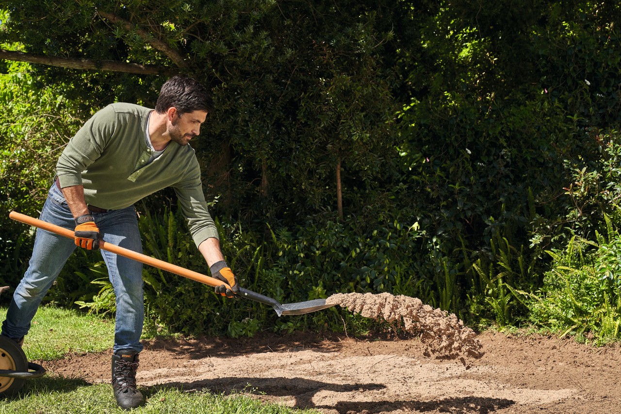 Ein Mann verteilt mit einem Spaten Bausand auf lehmigem Gartenboden, um ihn abzumagern