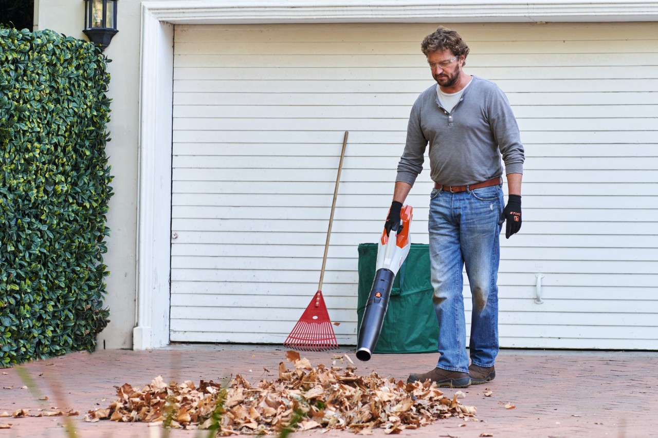 Mann pustet Herbstlaub in der Garageneinfahrt mit dem Blasgerät zu einem Haufen zusammen 