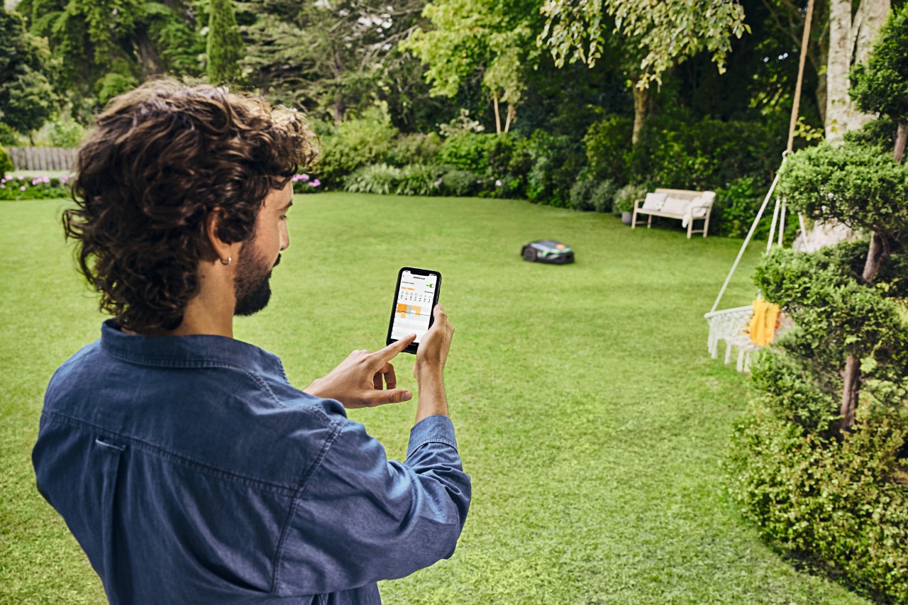 Mann steht mit Smartphone im Garten und steuert den Mähroboter