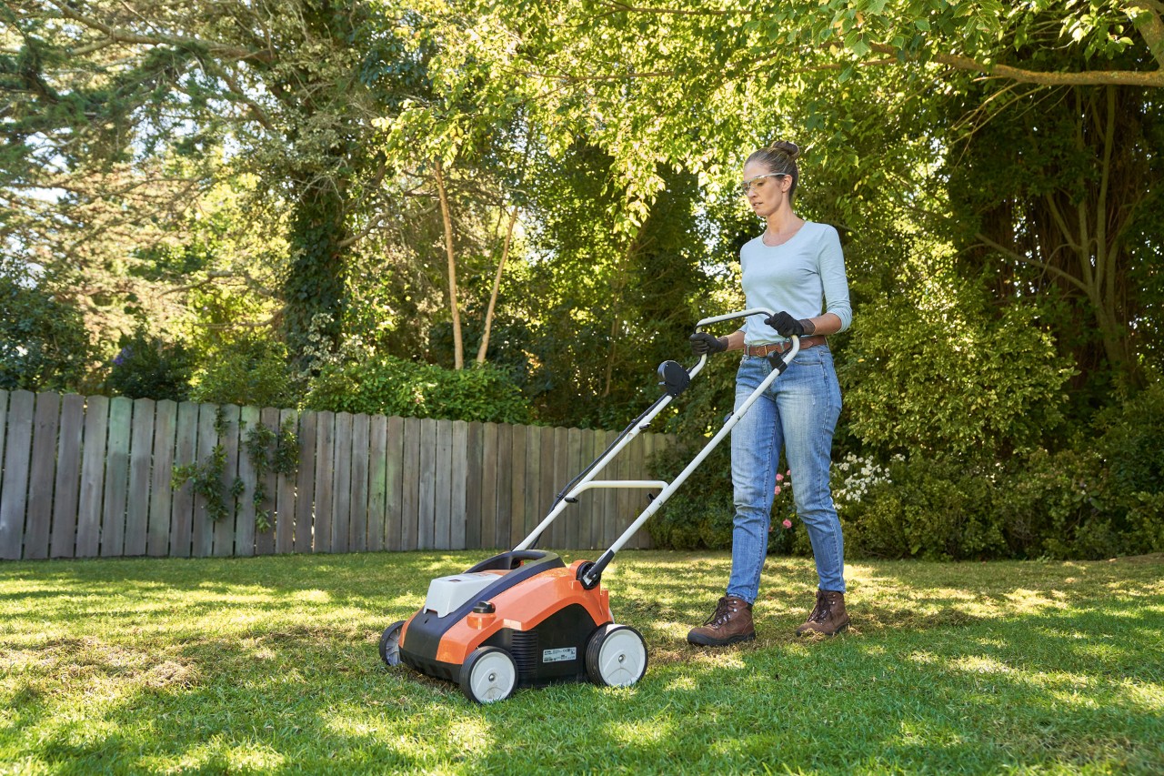 Frau lüftet Rasen mit STIHL Akku-Rasenlüfter RLA 240 im Garten, ausgestattet mit Schutzbrille und Arbeitshandschuhen.