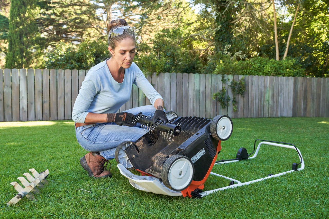 Frau mit Schutzbrille und Arbeitshandschuhen tauscht Vertikutiereinheit des STIHL Akku-Rasenlüfters RLA 240 aus