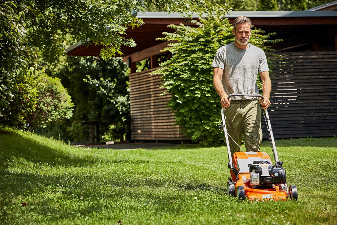 Mann beim richtig Rasenmähen mit einem STIHL Benzin-Rasenmäher RM 248 vor einer Gartenhütte