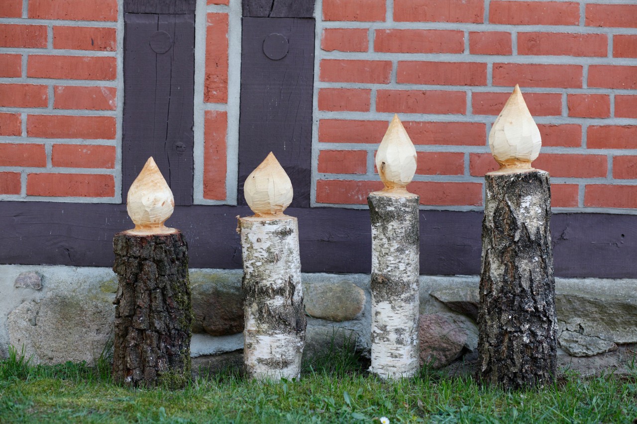 Vier Kerzen aus Holz zum Selbermachen stehen als Weihnachtsdeko auf einem Rasen vor einem Fachwerkhaus mit roten Backsteinen
