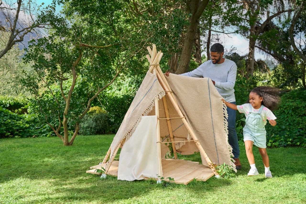 Mädchen und Mann bedecken den Rahmen des Gartenzelts für Kinder mit einer Decke oder einem Teppich.