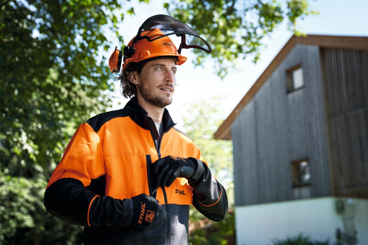 Mann mit orange-schwarzer STIHL Schutzausrüstung inklusive Helm und Handschuhe blickt zuversichtlich aus dem Bild, im Hintergrund ein Haus und Bäume 