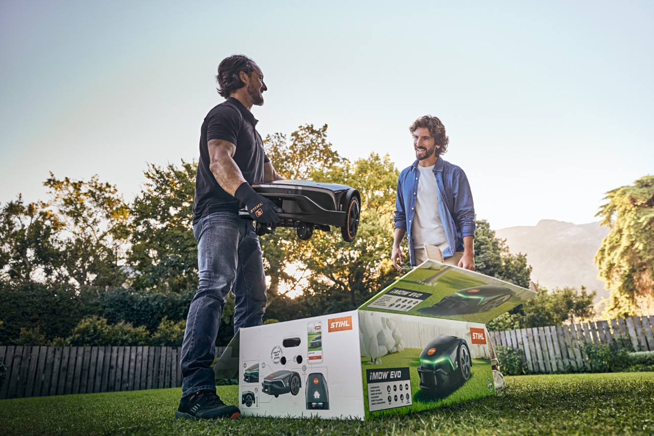 Ein STIHL Fachhändler steht mit einem iMOW® EVO in der Hand und dem iMOW® Karton davor im Garten eines Kunden und möchte beim installieren helfen